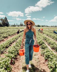 a woman standing in a field holding two buckets filled with plants and wearing a straw hat