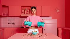 a woman standing in a pink kitchen holding two tea kettles over her stomach, with cereal scattered on the counter