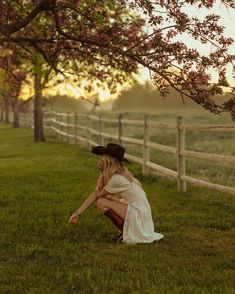 a woman in a white dress and cowboy hat kneeling under a tree