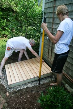 two people working on a wooden deck in front of a house with trees and bushes