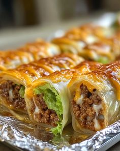 some food that is sitting on top of tin foil in the shape of burritos with meat, cheese and lettuce