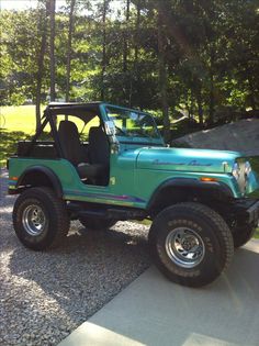 a green jeep parked on top of a gravel covered driveway next to trees and grass