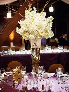 a tall vase filled with white flowers sitting on top of a purple table cloth covered table