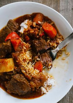 a white bowl filled with meat and rice on top of a wooden table next to a fork