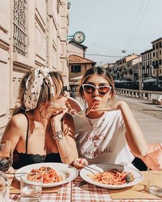 two women sitting at a table with plates of spaghetti in front of them, eating