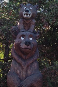 two brown bears sitting on top of each other in front of some trees and bushes