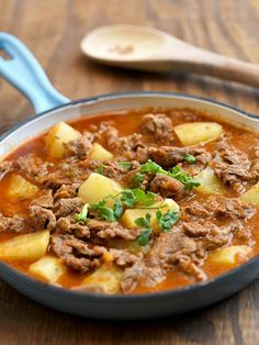 a stew with potatoes and meat in a blue pan on a wooden table next to a spoon