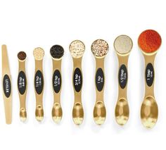 six spoons with different types of spices in them on a white surface, one is gold and the other has black lettering