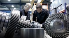 two men working on some metal parts in a factory with other workers looking at them