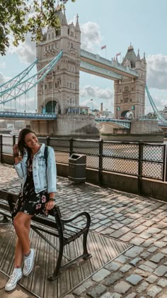 a woman is sitting on a bench in front of the tower bridge and looking up