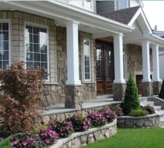 a stone house with white columns and flowers in the front yard on a sunny day