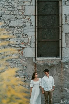 a man and woman standing next to each other in front of a stone wall with a window