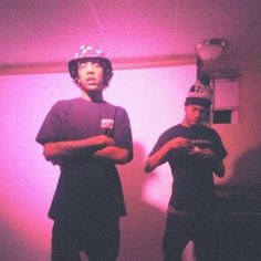 two young men standing next to each other in front of a pink light with their arms crossed