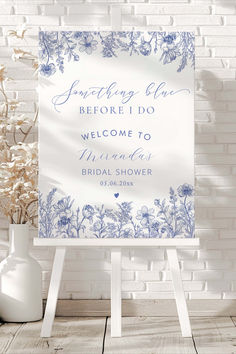 a welcome sign with blue flowers on it in front of a white brick wall and vase