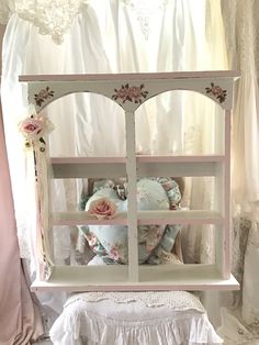 a white shelf with pink flowers on it in front of a window and curtains behind it