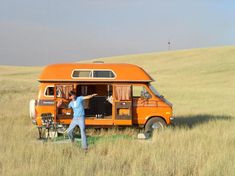 an orange van parked in the middle of a field with two people standing next to it