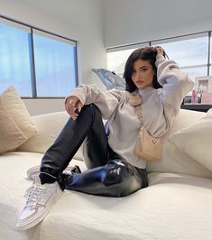 a woman sitting on top of a bed wearing black pants and white sweater with her hands behind her head