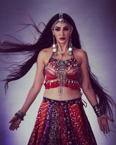 a woman with long black hair wearing a colorful dress and headdress, standing in front of a white background