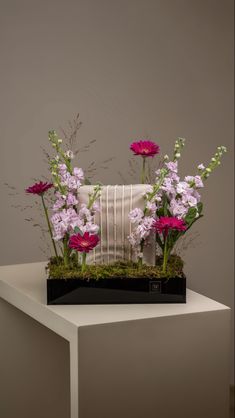 an arrangement of flowers and plants in a vase on top of a white table against a gray wall