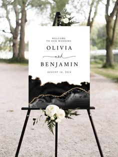 a welcome sign for an outdoor wedding with white flowers and greenery on the easel