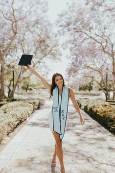 a woman in a graduation gown is holding up her cap and diploma while walking down a path