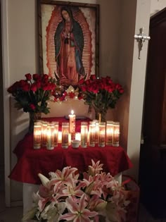 candles and flowers on a table in front of a virgin mary painting with red cloth