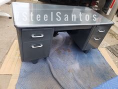 an office desk with steel santos on the bottom and drawers underneath it, sitting in front of a truck