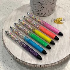 six pens are lined up on top of a wooden stand next to a cup filled with confetti and sprinkles