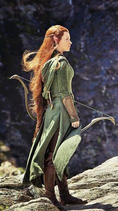 a woman with long red hair is standing on rocks and holding a bow in her hand