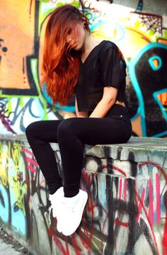 a woman with red hair is sitting on a wall covered in graffiti and has her hands behind her head