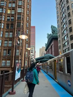 a woman with a backpack walking down a sidewalk next to a train on the tracks