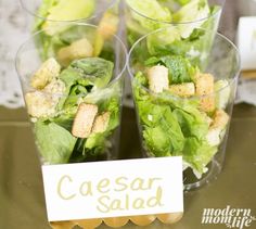 three glasses filled with salad sitting on top of a table next to a sign that says caesar salad