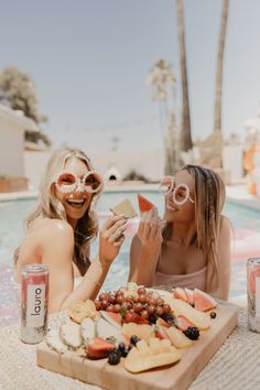 two beautiful women sitting next to each other in front of a swimming pool with food