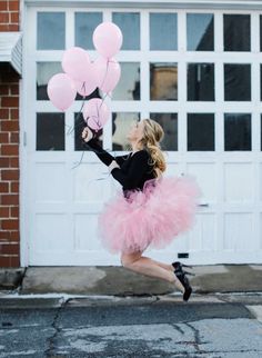 a woman in a pink tutu skirt and black top is jumping with some balloons