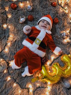 a baby dressed as santa clause laying on the floor next to some christmas decorations and lights