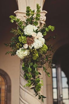 a vase with flowers and greenery hanging from the side of a pillar in front of a building