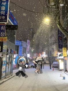 two people walking down a snowy street at night with snow falling on the ground and buildings in the background