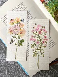 three cards with watercolor flowers on them sitting next to an open book and butterfly