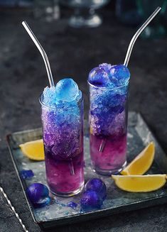 two glasses filled with purple and blue liquid sitting on top of a plate next to lemon wedges