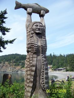 a carved wooden statue holding a large piece of wood in his hands and standing next to the water