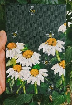 a person holding up a painting with white daisies and bees on it in front of green leaves