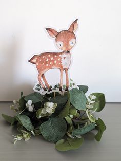 a paper deer is standing on top of some green leaves and white flowers in front of a white wall