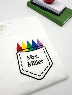 a t - shirt that says mr miller with crayons in the pocket and an apple next to it