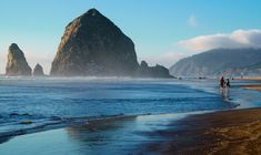 two people walking on the beach next to some big rock formations in the water and one person is carrying a surfboard