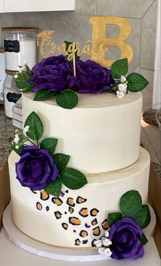 a three tiered cake with purple flowers and gold letters on top, sitting on a counter