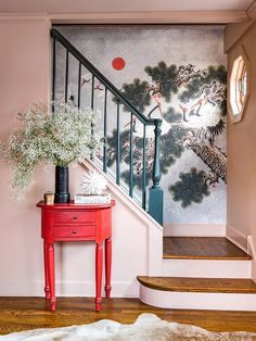 a red table sitting in front of a stair case next to a painting on the wall