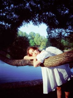 a woman in white dress leaning on a tree branch by the water's edge