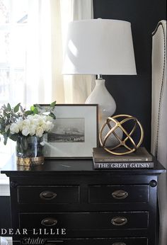 a black dresser with white flowers on top and a lamp in the corner next to it