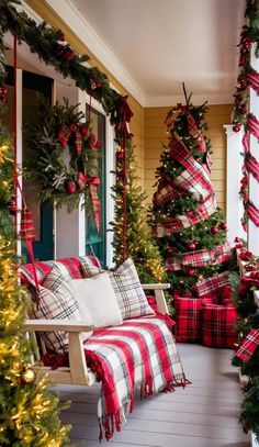 a porch decorated for christmas with red and white plaid pillows, garlands and trees