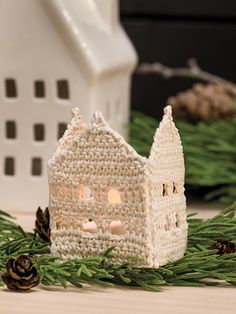 a small crocheted house next to pine cones and evergreen needles with a lit candle in the center